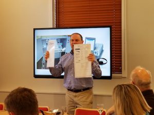 Dr. Lazar standing in front of an audience holding ballot forms with a presentation screen behind him.