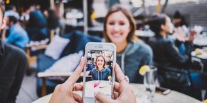 Friends sitting at a table in a restaurant. One friend is capturing an image of the other with a smart phone.